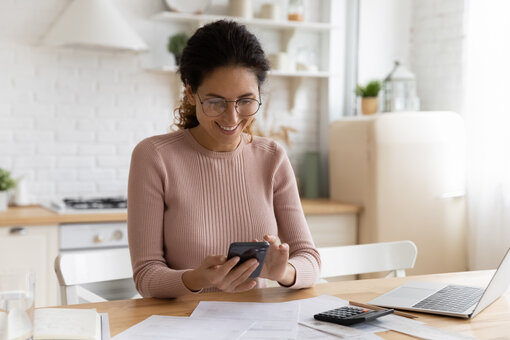 AdobeStock_459426517 Smiling young Caucasian woman manage household budget finances paying bills taxes on smartphone online. Happy female busy with documents calculate expenses expenditures on cellphone on internet.